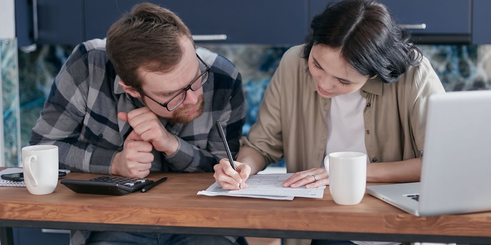 Young couple at kitchen table reviewing paperwork with a laptop and calculator, deciding how much house they can afford