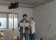Young couple planning a home renovation project with blueprints in a partially renovated room, considering how to fund the remodel