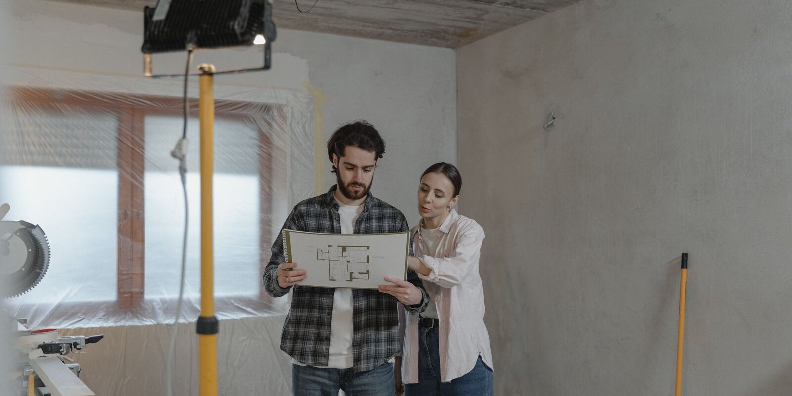 Young couple planning a home renovation project with blueprints in a partially renovated room, considering how to fund the remodel