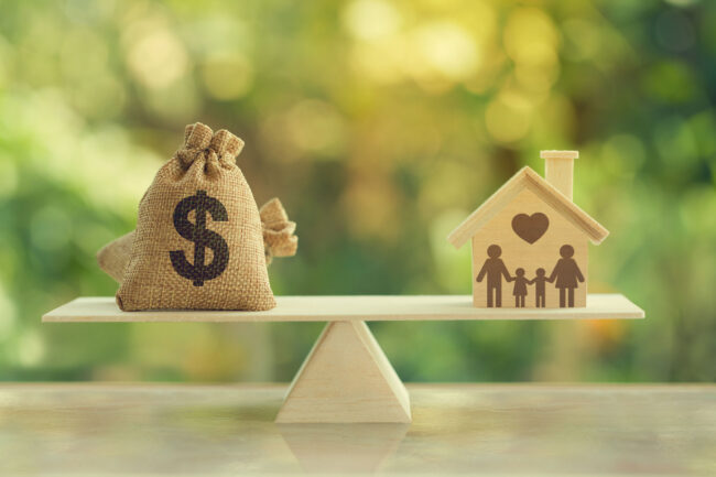 home-equity-loan-vs-cash-out-refinance-balance-scale-2026 Money bag on one side of a balance scale weighed against a wooden house with family silhouette on the other, representing the home equity loan vs cash-out refinance decision