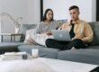 Young Asian couple sitting together on their sofa in a modern living room reviewing home options and mortgage rate math on a laptop, weighing whether to buy a home now or wait for mortgage rates to drop after the Federal Reserve September 2026 hawkish surprise