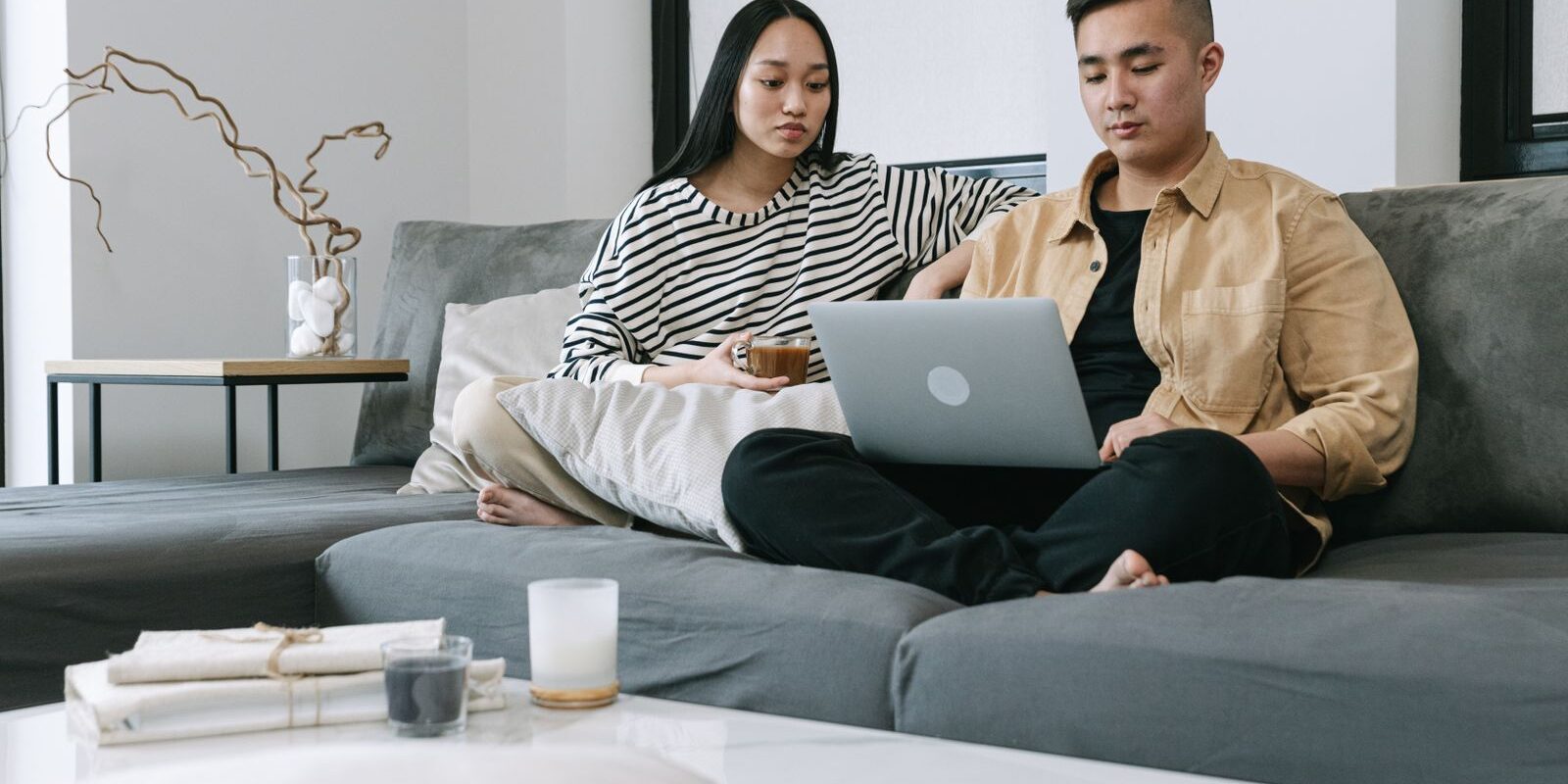 should-i-wait-buy-home-mortgage-rates-2026 Young Asian couple sitting together on their sofa in a modern living room reviewing home options and mortgage rate math on a laptop, weighing whether to buy a home now or wait for mortgage rates to drop after the Federal Reserve September 2026 hawkish surprise