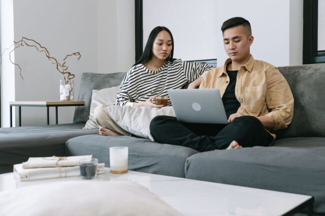 Young Asian couple sitting together on their sofa in a modern living room reviewing home options and mortgage rate math on a laptop, weighing whether to buy a home now or wait for mortgage rates to drop after the Federal Reserve September 2026 hawkish surprise