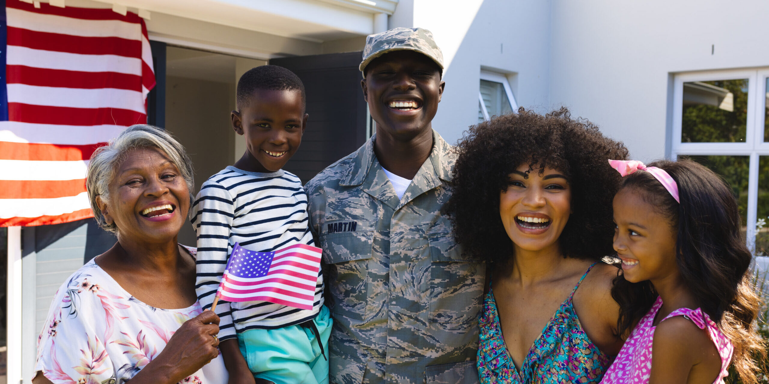 Portrait of happy multiracial army soldier in camouflage clothing with family standing in yard Multi-generational veteran family celebrating in front of their home with American flag draped from the porch, representing the veteran families OnPoint serves with the VA IRRRL refinance program