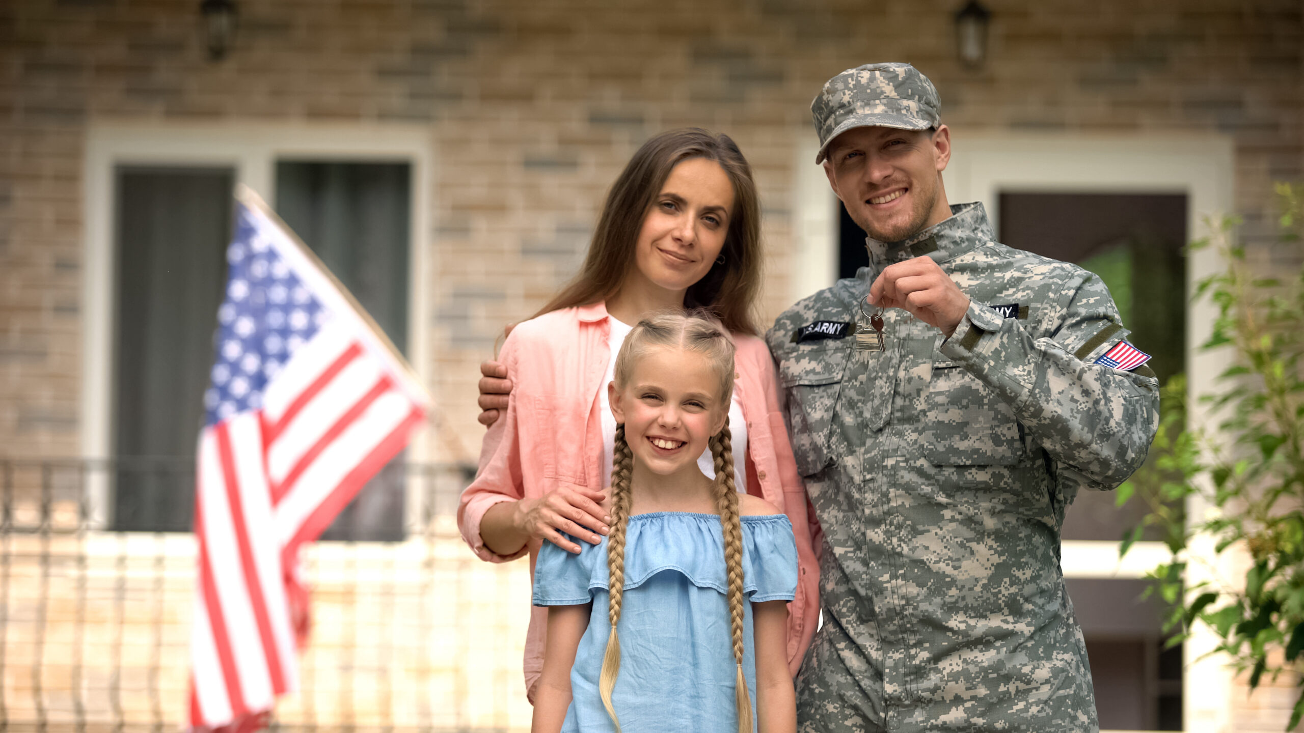 US Army veteran holding the keys to his new home with wife and daughter, American flag in front of their house