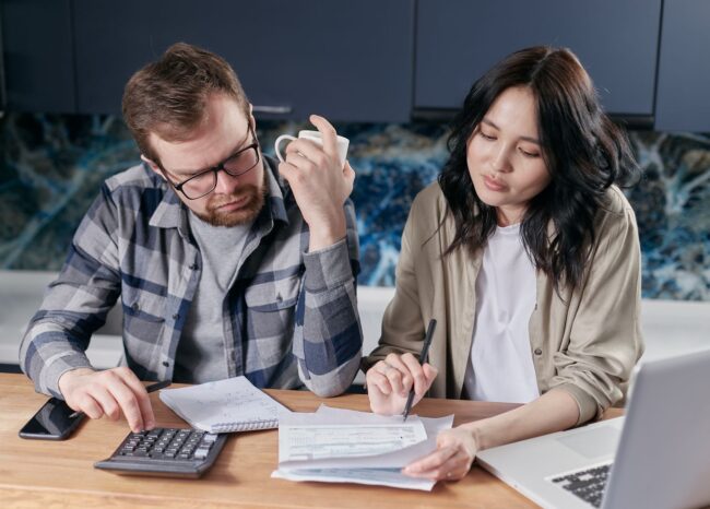 Couple at their kitchen table running the household income and debt math with a calculator and laptop, determining what mortgage payment they can afford on a $500K, $750K, or $1M home in Orange County in October 2026